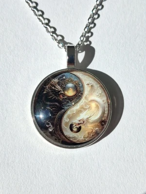 Yin Yang halsband med drakmotiv - Unikt halsband med runt hänge i silverfärg, prytt med ett detaljerat Yin Yang-motiv i 2D där två drakar möts i svart och vitt. Kedjan är i metall och hänget har en cool fantasykänsla. Materialet rostfritt stål 