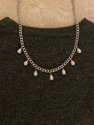 Silverfärgat halsband med pärldroppar - Kedjehalsband i silverfärg med sju droppformade pärlor som hänger längs kedjan. Halsbandet har en chunky kedja och pärlorna ger en elegant touch. Perfekt för dig som gillar stilrena accessoarer med lite extra detalj.