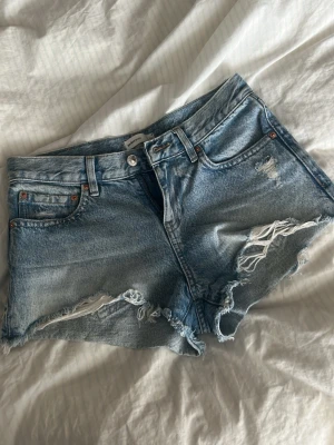 Blå slitna jeansshorts med fransar - Säljer ett par ljusblå jeansshorts med råa, fransiga kanter och slitningar framtill. Shortsen har klassisk femficksmodell, knappgylf och bälteshällor. Perfekta för en avslappnad och trendig sommarlook.