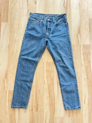 Levi's 501 blå raka jeans - Klassiska Levi's 501 jeans i ljusblå denim med rak passform och knappgylf. Jeansen har fem fickor, röd Levi's-tag på bakfickan och läderpatch i midjan. Perfekta för en avslappnad och tidlös stil. Storlek W29 L30