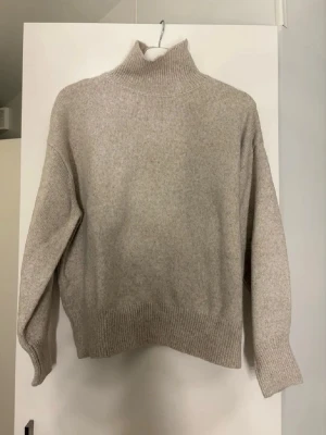 Beige polotröja i ullmix - Mysig beige stickad polotröja med hög krage och avslappnad passform. Tröjan har ribbade muddar vid ärmslut och nederkant, vilket ger en snygg och bekväm look. Perfekt för kalla dagar och enkel att matcha med olika outfits.