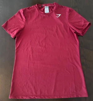 Gymshark t-shirt  - Snygg röd t-shirt från Gymshark med klassisk passform och diskret logga på bröstet. Tillverkad i mjuk polyester som andas, perfekt för träning eller chill. Rund halsringning och korta ärmar ger en clean look.