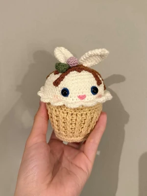 Virkad cupcake-kanin amigurumi - Söt handvirkad amigurumi i form av en cupcake med kaninöron. Cupcaken är beige och vit med chokladbruna detaljer, blå säkerhetsögon, rosa nos och små virkade dekorationer på toppen. Tillverkad i mjukt garn, perfekt som dekoration eller present.