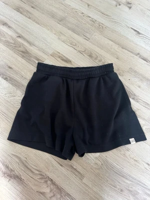 Svarta shorts  - Säljer ett par svarta shorts från Dcore med elastisk midja och avslappnad passform. Shortsen har en liten logotyp på vänstra benet och är tillverkade i mjukt bomullsmaterial, perfekta för träning eller chill.