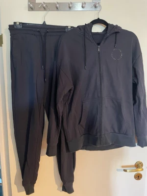 Svarta mjukisbyxor från MM Sports XS med kofta - Svarta mjukisbyxor från MM Sports i storlek XS. Byxorna har dragsko i midjan och muddar vid bensluten. Perfekta för chill eller träning, tillverkade i mjukt bomullsmaterial för extra komfort.plus matchande kofta 