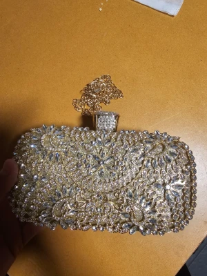 Guldig kuvertväska med strassdetaljer - Säljer en superglittrig kuvertväska i guld med massor av strass och gnistrande stenar i blommönster. Väskan har en hård rektangulär form och en dekorativ kedja i guld som axelband. Perfekt för dig som vill sticka ut och älskar bling!