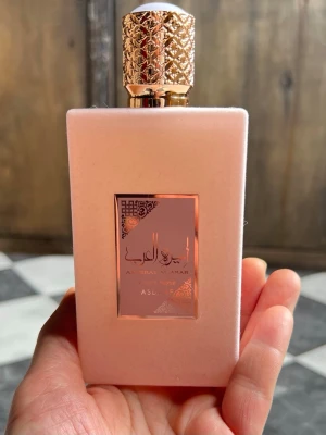 Asdaaf Ameerat Al Arab Privé Rose parfym 100ml - Lyxig parfymflaska i matt ljusrosa med gulddetaljer och elegant mönstrad kork. Märket Asdaaf och doften Privé Rose ger en exklusiv känsla. Flaskan har en rektangulär form och en dekorativ etikett i guld med arabiska inslag. Bara testad. Ingen låda. 