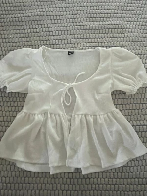 Vit blus med puffärm och knyt - Supersöt vit blus med puffärmar och knyt framtill. Blusen har en luftig och flowy passform med volang nedtill och rundad urringning. Perfekt för en trendig och feminin look.