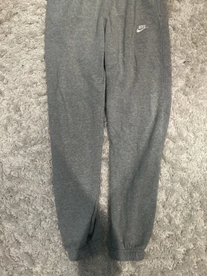 Gråa mjukisbyxor från Nike - Säljer ett par gråa mjukisbyxor från Nike med resår i midjan och vid bensluten. Byxorna har en klassisk passform och är tillverkade i mjukt sweatshirtmaterial. Nike-logga broderad på vänster lår. Perfekta för chill dagar eller träning.