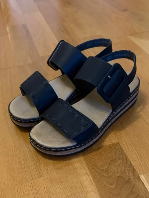 Svarta sandaler från Rieker - Snygga svarta sandaler från Rieker med breda remmar och stor spänndetalj. Sandalerna har en bekväm, ljus innersula och en platåsula med kontrastsömmar. Tillverkade i syntetmaterial och passar perfekt till sommarens outfits.