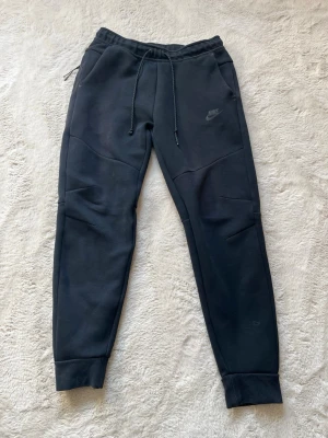 Nike tech byxor - Säljer ett par svarta mjukisbyxor från Nike med dragsko i midjan och ribbade muddar vid bensluten. Byxorna har en slim fit passform och diskret Nike-logga på vänster lår. Perfekta för chill eller träning.