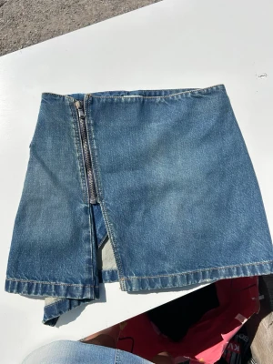 Blå jeanskjol från Eytys med dragkedja - Säljer en blå jeanskjol från Eytys med markerad dragkedja framtill och cool asymmetrisk slits. Kjolen har hög midja och tydliga sömmar, samt Eytys-logga broderad baktill. Perfekt för dig som gillar streetwear och denim.