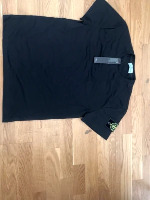 Svart Stone Island t-shirt i bomull - Svart t-shirt från Stone Island med klassisk logopatch på vänster ärm. Tillverkad i mjuk bomull och har en enkel, stilren design med rund hals och korta ärmar. Perfekt för dig som gillar streetwear och vill ha något stilrent.