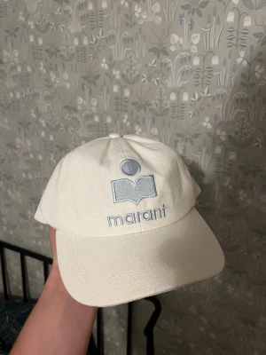 Vit keps från Isabel Marant - Snygg vit keps från Isabel Marant med ljusblå broderad logga framtill. Klassisk böjd skärm och justerbar baksida. Tillverkad i ett tåligt bomullstyg som ger en avslappnad och trendig look.