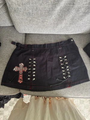 Vintage goth mini-kjol - Vintage mini-kjol från tidigt 2000 talet från Brittiska märket ”Dead Threads”. Kjolen har röda sömmar, snörningsdetaljer framtill och en dekorerad korsapplikation med nitar. Trots sin ålder så är den i jättebra skick, endast några lösa trådar. Nypris 2003 var 1500kr