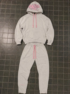 Äkta Gråa syna world tracksuit  - Mysiga grå syna world tracksuit från synaworldwideuk.com storlek m med breda rosa dragsnören och matchande hoodie. Hoodien har graffiti-inspirerat tryck på luvan och stor ficka framtill. Perfekt för chill dagar. Materialet är mjukt och skönt.