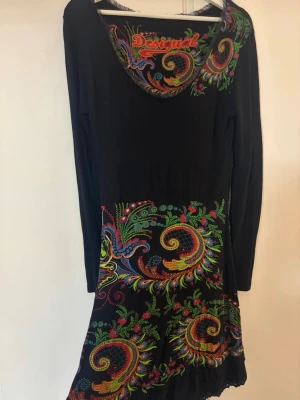 Svart Desigual klänning med färgglatt mönster - Unik svart klänning från Desigual med lång ärm och färgglada broderade detaljer i paisley- och blommönster. Klänningen har rund halsringning och färgstarka inslag i rött, grönt, blått och gult. Perfekt för dig som gillar statement-plagg.
