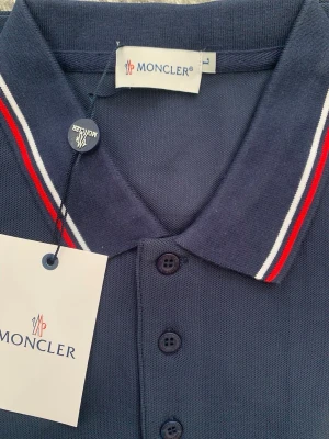 Mörkblå Moncler pikéskjorta - Snygg mörkblå pikéskjorta från Moncler med klassisk krage och knappar framtill. Kragen har röda och vita ränder som ger en sportig touch. Bröstet pryds av Monclers ikoniska logotyp. Tillverkad i bomull för en bekväm känsla.