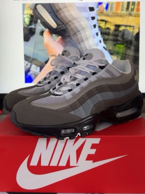 Nike Air Max 95 grå sneakers - Nike Air Max 95 sneakers i olika nyanser av grått med vita och svarta detaljer. Ovandelen är i mesh och mocka för en sportig look, och sulan har synliga Air-enheter för extra komfort. Klassisk snörning och ikonisk design som sticker ut.