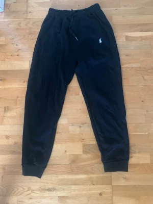 Svarta mjukisbyxor Polo Ralph Lauren - Svarta mjukisbyxor från Polo Ralph Lauren med dragsko i midjan och resår vid benslut. Klassisk vit logga broderad på vänster sida. Sköna och stilrena byxor i mjukt material, perfekta för chill dagar. Passformen är relaxed och modellen har fickor på sidorna.
