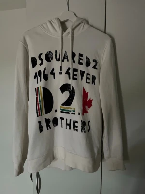 Vit hoodie från Dsquared2 - Säljer en vit hoodie från Dsquared2 med tryck i svart, rött och flera färger framtill. Texten 'DSQUARED2 1964 4EVER BROTHERS' och en röd lönnlövsgrafik ger en cool vibe. Hoodien har dragsko i huvan och är tillverkad i mjukt material.