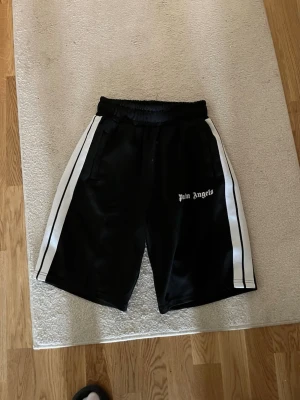Palm Angels svarta shorts med vita ränder - Svarta shorts från Palm Angels med vita ränder längs sidorna och logga på vänster ben. De har elastisk midja och sportig look, perfekta för en avslappnad stil. Materialet känns glansigt och lätt, troligen polyester.