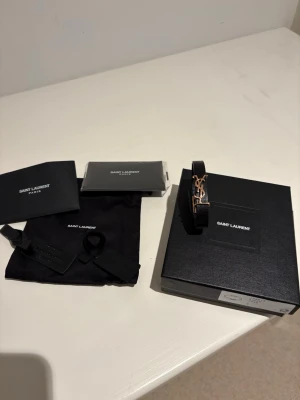 Saint Laurent svart läderarmband - Snyggt svart armband från Saint Laurent i äkta läder med ikoniskt YSL-spänne i guld. Smal och stilren design som ger en lyxig touch till din outfit. Kommer med originalask, dustbag och certifikat.