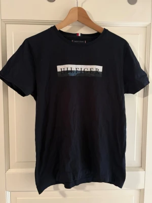 Tommy Hilfiger t-shirt - Snygg svart t-shirt från Tommy Hilfiger med klassisk logga framtill i vitt och grått. T-shirten har rund halsringning och korta ärmar. Tillverkad i mjuk bomull för en bekväm känsla. Perfekt till jeans eller shorts.