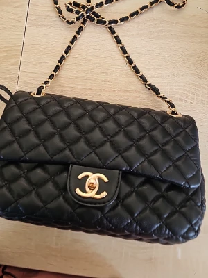  svart quiltad axelväska - Snygg svart axelväska i quiltat läder med klassiskt guldigt CC-spänne. Kedjerem med insytt läder ger en lyxig känsla. Perfekt för dig som vill ha en ikonisk och tidlös accessoar med exklusiv vibe.