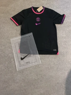 Svart PSG x Nike t-shirt med rosa detaljer - Svart t-shirt från Nike i samarbete med Paris Saint-Germain. Tröjan har rosa och lila detaljer vid krage och ärmslut samt PSG-logga och Nike swoosh i rosa framtill. Perfekt för fotboll eller streetwear. Materialet är lätt och andas.