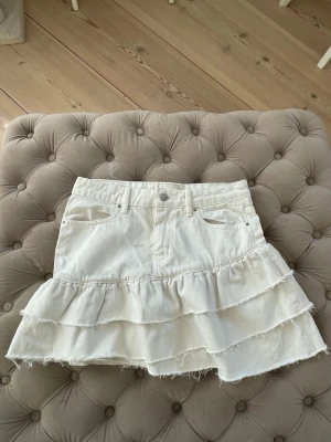 Vit volangkjol i denim - Supersöt vit kortkjol i denim med volanger och råa kanter. Kjolen har klassisk femficksdesign, bälteshällor och knäppning fram. 