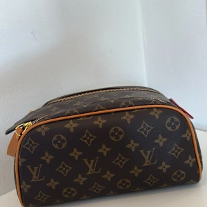 Nyskick brun modern och trendig monogram necessär - Säljer en brun necessär i nyskick med klassiskt monogrammönster i guld och beige. Väskan har guldfärgade dragkedjor, handtag med röda detaljer. Perfekt för att organisera dina småsaker.