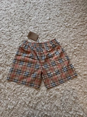Rutig shorts från Burberry - Klassiska rutiga shorts från Burberry i beige, svart, vit och röd. De har elastisk midja och det ikoniska Burberry-mönstret. Perfekta för dig som vill ha en tidlös och trendig look. Helt nya med prislapp