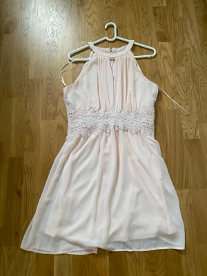 Rosa festklänning från Vila (Vimilina) – nyskick! 🌸 - Jättefin ljusrosa (Misty Rose) festklänning från Vila (Vimilina). Elegant halterneck med nyckelhålsdetalj fram samt knapp och dold dragkedja bak. Midjan är elastisk med vackra broderade spetsdetaljer som ger en supersmickrande figur. Perfekt för sommarfest, bröllopsgäst, dop eller examen! 🌸• Storlek: L (ca 40/42)• Skick: Toppskick, använd 1 gång!• Material: Chiffong med mjukt foder (ej genomskinlig)• Mått: Armhåla-armhåla 48 cm, längd 94 cm (knälång)