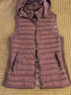 Ljuslila dunväst med huva - Snygg ljuslila dunväst med avtagbar huva och dragkedja framtill. Västen har två fickor med dragkedjor och dekorativa ringar. Quiltad design och mjukt material på sidorna ger en modern look. Perfekt för lager-på-lager-stil.