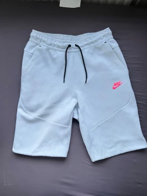Vita Nike shorts med snörning - Snygga vita shorts från Nike med svart snörning i midjan och rosa Nike-logga på ena benet. Shortsen har fickor fram och en bakficka med dragkedja. Perfekta för sommaren och chill dagar.