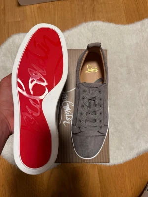 Gråa Christian Louboutin sneakers - Snygga gråa sneakers från Christian Louboutin med klassisk röd sula och vita kanter. Skorna är låga och har ovandel i mocka med matchande gråa skosnören. Perfekta för dig som vill ha något exklusivt och stilrent.