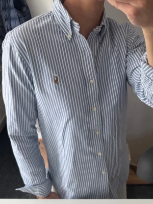 Blårandig skjorta från Ralph Lauren - Klassisk blårandig skjorta från Ralph Lauren med button-down krage och broderad logga på bröstet. Skjortan har långa ärmar, vita knappar och är tillverkad i mjuk bomull. Perfekt för en clean och stilren look.
