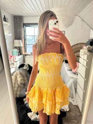 Gul smockad klänning Design By Amor - Supersöt kort klänning från Design By Amor i gul och vit med smockad överdel och volangig kjol. Klänningen är ärmlös och har ett blommigt broderat mönster, perfekt för varma dagar. Materialet känns lätt och luftigt.