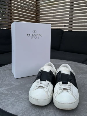 Valentino Garavani vita sneakers - Snygga vita sneakers från Valentino Garavani med svart läderdetalj över snörningen och ikoniska nitar baktill. Skorna har en klassisk låg siluett och är tillverkade i läder med vita snören och gummisula. Perfekta för dig som gillar exklusiv streetstyle. Även inkluderas låda.