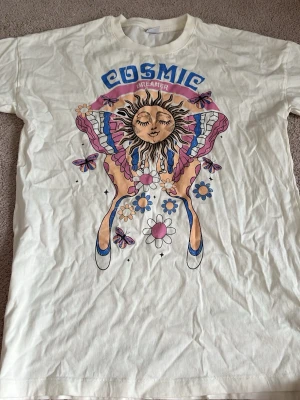 Vit Cosmic Dreamer t-shirt med tryck - Säljer en vit t-shirt med färgglatt tryck av en sol, blommor och fjärilar samt texten 'Cosmic Dreamer' i blått och rosa. T-shirten har rund hals och korta ärmar, perfekt för dig som gillar unika och kreativa motiv.