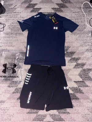 Under Armour träningsset marinblå - Snyggt träningsset från Under Armour med en marinblå t-shirt och matchande shorts. T-shirten har logga på bröstet och axeln, och shortsen har dragsko i midjan samt tryckta detaljer och logga. Perfekt för gymmet eller löprundan.