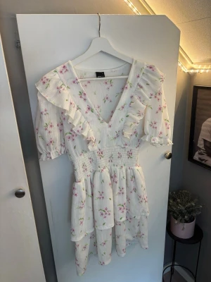 Vit blommig volangklänning Gina Tricot - Supersöt vit kortklänning från Gina Tricot med rosa blommigt mönster, volangdetaljer och v-ringning. Klänningen har korta ärmar, smock i midjan och ett luftigt, flowigt tyg i bomull. Perfekt för sommarens alla tillfällen!