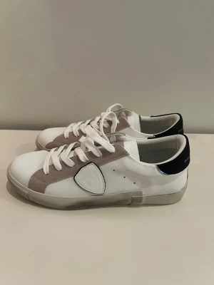 Philippe Model vita sneakers - Stilrena vita sneakers från Philippe Model med detaljer i beige mocka och svart hälparti. Klassisk låg modell med vita skosnören och diskret logga på sidan. Ovandelen är i läder och mocka, sulan är vit med lätt slitage-look. Perfekta för en trendig vardagsstil.