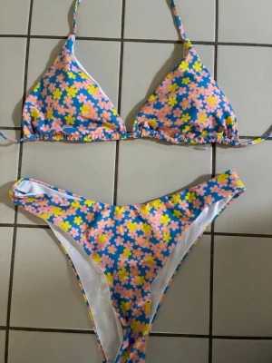 Blommig trekantsbikini i blått och rosa och gult - Färgglad trekantsbikini med blå botten och blommigt mönster i rosa och gult. Klassisk modell som knyts både i nacke och rygg. Endast använd vid 1 tillfälle! Perfekt för strandhäng och sommarvibbar!☀️