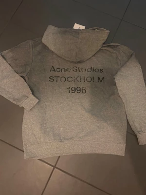 Grå hoodie Acne Studios Stockholm - Snygg grå hoodie från Acne Studios med tryck på ryggen där det står 'Acne Studios STOCKHOLM 1996'. Klassisk passform med huva och ribbade muddar. Tillverkad i mjukt bomullsmaterial som känns skönt mot huden. Perfekt för en avslappnad streetwear-look.