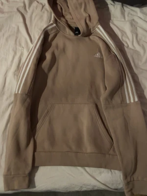 Beige hoodie från Adidas med huva - Snygg beige hoodie från Adidas med klassiska vita ränder längs ärmarna och liten logga på bröstet. Hoodien har huva och en stor magficka framtill. Tillverkad i mjukt material, perfekt för en avslappnad stil.