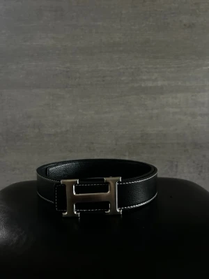 Hermess bälte - Snyggt svart skärp med klassiskt H-format metallspänne. Skärpet är gjort i skinn och har vita kontrastsömmar längs kanterna som ger en exklusiv känsla. Perfekt accessoar för att lyfta vilken outfit som helst.