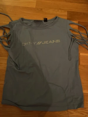 Grå topp med snörning från DKNY Jeans - Cool grå topp från DKNY Jeans med snörningsdetaljer längs båda ärmarna och tryckt logga framtill. T-shirten har rund halsringning och är tillverkad i mjuk bomull, perfekt för en avslappnad och trendig look.