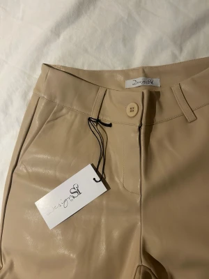 Beige skinnbyxor från Design S - Snygga beigea skinnbyxor från Design S med raka ben och hög midja. Byxorna har bälteshällor, knappstängning och fickor fram. Materialet är glansigt och ger en trendig look som passar perfekt till olika outfits.